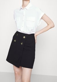 Bílá košile s krátkým rukávem a dvěma kapsami, doplněná černou zavinovací sukní s dvojitými zlatými knoflíky na přední straně.