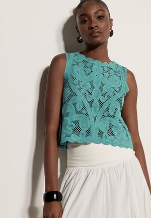 Mujer que lleva un top sin mangas de crochet color verde azulado con patrones florales y una falda blanca de talle alto, complementada con una pulsera negra y pendientes de aro.