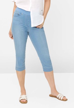 Kvinde iført lyseblå korte skinny jeans med elastisk talje, hvide åbne tå-sandaler og en hvid skjorte med knapper.