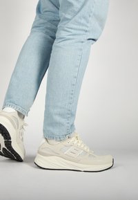 Svetle béžové sportovní boty mají svršek z prodyšné síťoviny a semiše, kulatou špičku, tónové tkaničky a černou gumovou podrážku, doplněné o světle modré džíny.