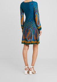 Femme portant une robe à manches longues de couleur sarcelle avec des motifs cachemire abstraits orange, violet et bleu, associée à des sandales à talons carrés blanches.