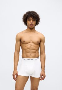 White cotton boxer briefs with a black Calvin Klein logo waistband, fitted style, and a smooth texture, model standing upright.