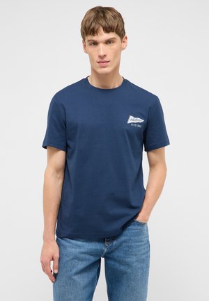 Young man wearing navy blue Mustang Jeans Company t-shirt and blue jeans, standing with one hand in pocket against plain background.