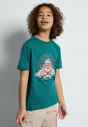 Camiseta de algodón color azul verdoso con un gráfico de una calavera usando gafas de sol y un sombrero, acompañado de texto, combinada con pantalones cortos cargo beige.