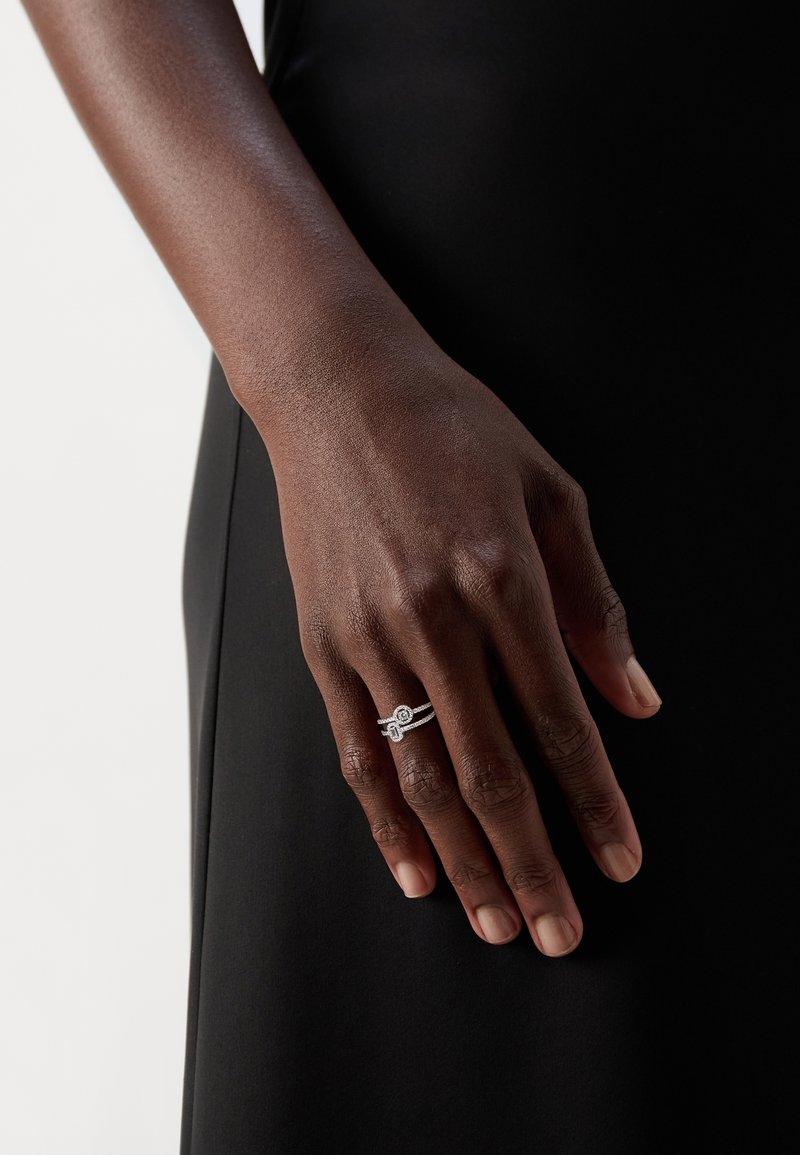 Main à la peau foncée reposant sur un tissu noir, portant une bague double en argent avec de petits diamants à l'annulaire.