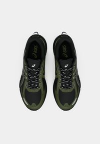 Des chaussures de sport noires et vert olive présentent un dessus en tissu texturé, des accents réfléchissants et des lacets gris, avec une semelle rembourrée pour un meilleur soutien.