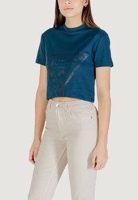 T-shirt cropped color teal con un design a triangolo stampato scuro. Abbinato a jeans beige aderenti, che mettono in evidenza una texture liscia e un taglio semplice.