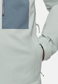 Veste imperméable bleu clair avec un panneau rectangulaire gris, dotée de cordons de serrage ajustables et d'une poche latérale. Texture lisse, coupe ajustée.