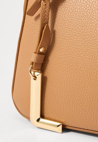 Borsa a tracolla in pelle color tan con superficie testurizzata, dettagli in hardware dorato e un charm rettangolare minimalista attaccato alla tracolla.