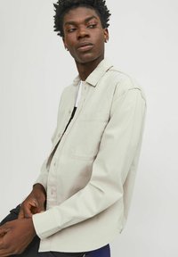 Chemise beige clair à boutons, à manches longues, avec col, poche poitrine unique et texture lisse. Présentée sur un modèle en pose assise.