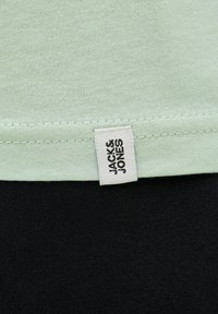 Mintgrøn stof med en glat overflade og en sort nederste del. Har et hvidt mærke med "JACK & JONES" broderet på sømmen.