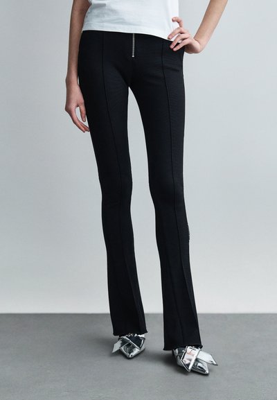 Pantalon noir évasé avec une fermeture éclair à la taille, présentant des coutures contrastées et une texture lisse, associé à des chaussures pointues argentées et brillantes.