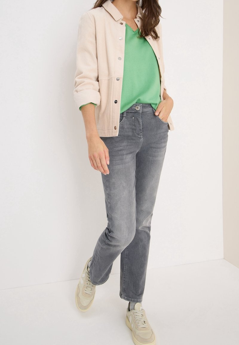 Femme portant une veste en velours côtelé beige, un haut vert, un jean gris et des baskets beiges, debout avec une main dans la poche.