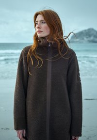 Braune Fleecejacke mit hohem Kragen und Reißverschluss. Verfügt über ein strukturiertes Material und lange Ärmel. Befindet sich in einer küstlichen Outdoor-Umgebung.