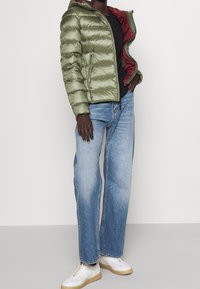 Oliwkowa kurtka puchowa z kapturem, z zamkiem błyskawicznym i bordowym wykończeniem. Połączona z jasnoniebieskimi dżinsami o prostym kroju i białymi sneakersami.