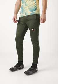 Zaļas sporta bikses ar pieguļošu dizainu, kurām ir rozā Puma logos. Valkātas ar melnajām futbola kurpēm un kreklu ar abstraktu rakstu.