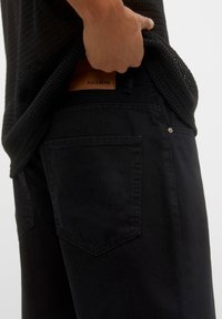 Pantalones de mezclilla negra con un acabado texturizado, presentando una etiqueta de cuero en la cintura y bolsillos traseros con doble costura.