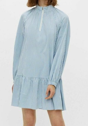 Femme portant une robe à manches longues en vichy bleu et blanc avec un col montant et une fermeture éclair à l'avant, debout devant un fond uni.