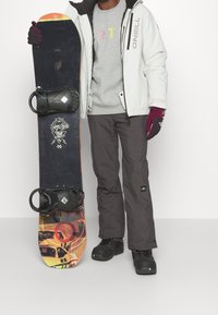 Snowboard con un top nero, design grafico multicolore sulla base e attacchi neri. Modello che indossa una giacca bianca e pantaloni grigi.