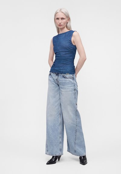 Femme aux cheveux blond clair portant un haut sans manches bleu marine, un jean large bleu clair et des bottes noires à talons, debout devant un fond blanc uni.