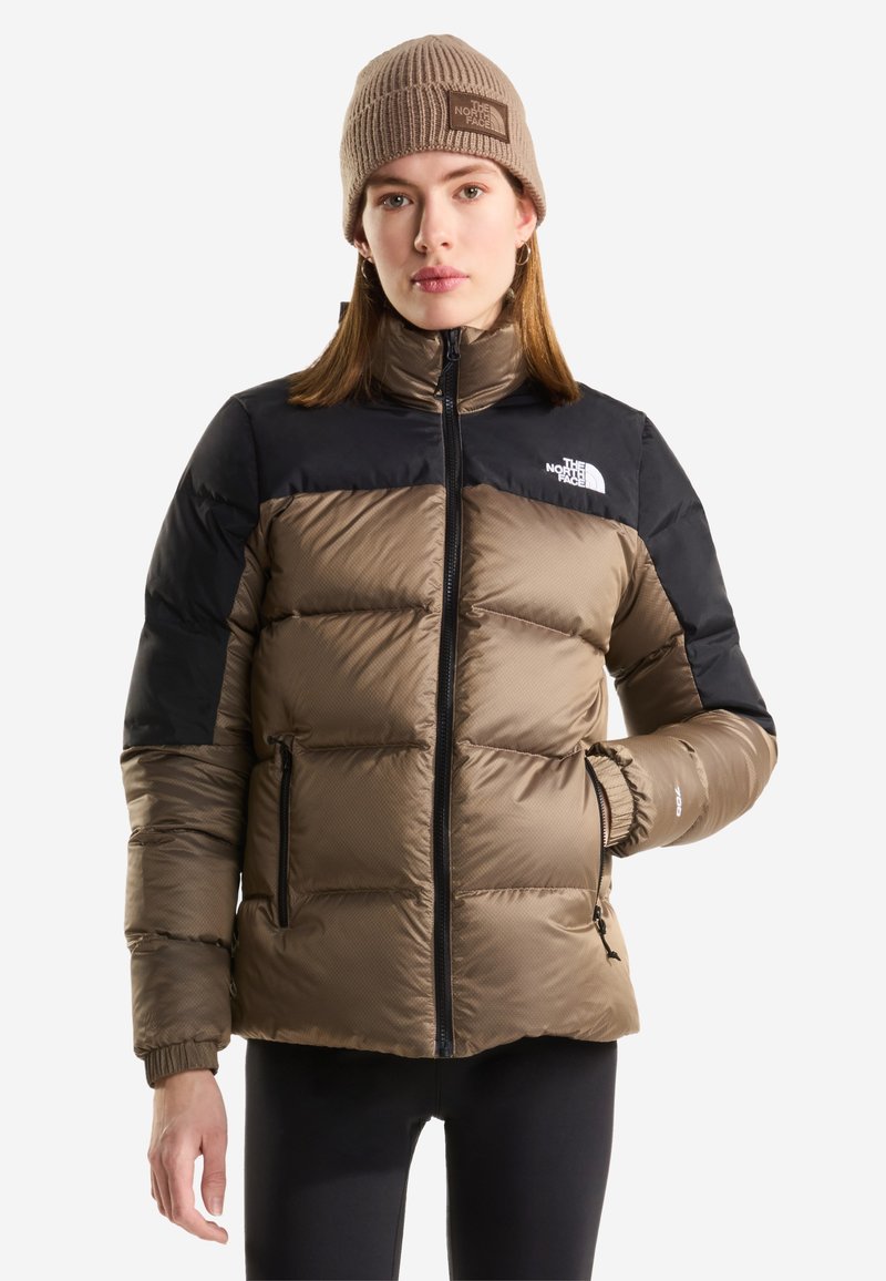 Γυναίκα με καφέ και μαύρο μπουφάν North Face και καφέ πλεκτό σκουφάκι, στέκεται μπροστά σε απλό λευκό φόντο.