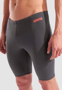 Graue Badeshorts mit enger Passform, aus glattem Material gefertigt. Sie verfügen über ein orangefarbenes Logo am Bund und einfache, gestickte Nähte.