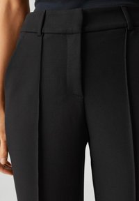 Zwarte op maat gemaakte broek heeft een fijne textuur, een middelhoge tailleband en voorplooien, met een strakke en gepolijste afwerking.