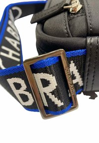 Bolsa de tela negra con una correa a rayas azul y negra, que presenta letras en blanco en negrita y una hebilla de metal plateada.