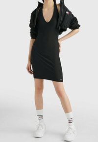 Svart ribbad halternklänning med djup V-ringning, tillsammans med en kort svart jacka och vita sneakers med logostrumpor. Minimalistisk design.