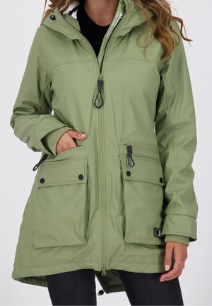 Femme portant une parka à capuche vert clair avec une fermeture éclair frontale et plusieurs poches, une main dans la poche, sur fond blanc.
