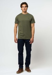 Άνδρας που στέκεται φορώντας ελαι πράσινο t-shirt, σκουρό μπλε τζιν και καφέ δερμάτινα μποτάκια μπροστά σε απλό φωτεινό φόντο.