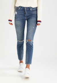 Vaqueros ajustados azules de denim con desgaste, rodillas rasgadas y un acabado desgastado. Combinados con zapatillas blancas y un suéter crema.