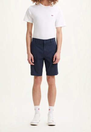 Osoba ubrana w białą koszulkę Levi's, granatowe szorty, białe skarpetki Levi's oraz białe trampki, stojąca na jasnym tle.