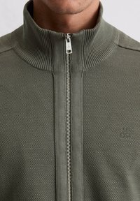 Donkergroen ritsvest met hoge kraag, geribde textuur en een klein logo op de borst, met een zilveren ritsdetail.
