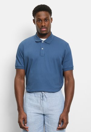 Mann trägt ein blaues kurzärmeliges Poloshirt und hellblaue Hose mit Kordelzug, steht vor einem schlichten weißen Hintergrund.