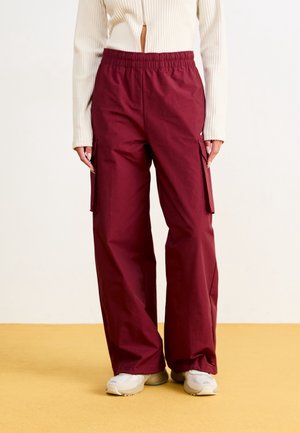 Persoon draagt bordeauxrode cargobroek met hoge taille en losse pasvorm en witte sneakers, gecombineerd met een crème ribgebreide longsleeve.