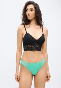 Γυναίκα με σγουρά μαλλιά, φοράει μαύρο δαντελένιο bralette και σλιπ μέντας με δαντέλα, κοιτάζοντας προς τα αριστερά της σε απλό φόντο.