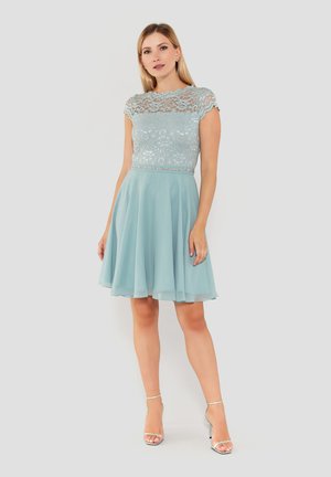 Frau steht in einem hellblauen Spitzenoberteil und einem Chiffonrock-Kleid mit silbernen High Heels vor einem einfarbigen Hintergrund.