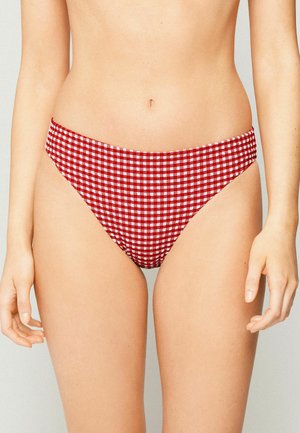 Femme portant un bas de bikini à carreaux rouges et blancs, raccourci à la taille, bras détendus sur les côtés.