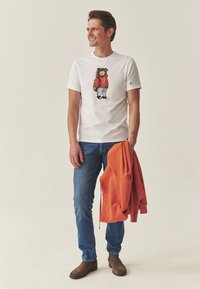 Fehér póló, melyen egy medve grafikája látható piros dzsekiben, kék farmerral és barna csizmával. Narancssárga kapucnis pulóver lóg a karon. Semleges háttér.