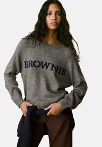 Jersey gris de punto con el texto "BROWNIE" en azul marino, cuello redondo, puños de canalé y corte relajado, combinado con pantalones marrones y un suéter oscuro.