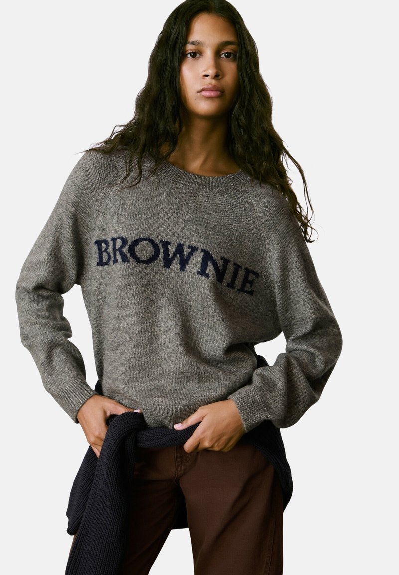 Jersey gris de punto con el texto "BROWNIE" en azul marino, cuello redondo, puños de canalé y corte relajado, combinado con pantalones marrones y un suéter oscuro.