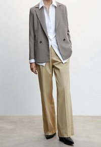 Blazer à double boutonnage en pied-de-poule gris, associé à une chemise blanche et un pantalon large crème, complété par des chaussures noires à bouts pointus.