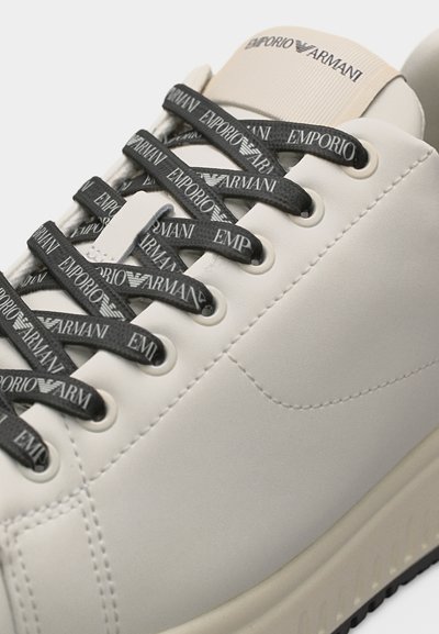 Λευκό αθλητικό παπούτσι με κορδόνια μαύρου χρώματος με το λογότυπο Emporio Armani και ετικέτα στη γλώσσα σε μπεζ απόχρωση, εμφανίζοντας την άνω πλευρά και λεπτομέρειες ραφής.