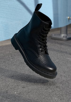 Schwarzer, strukturierter Leder-Schnürstiefel, schwebt mittig in der Luft über einer Asphaltfläche nahe einer hellblauen Wand und einem Metallgeländerpfosten.