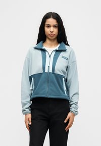 Νεαρή γυναίκα φοράει μπλε και ανοιχτό μπλε φλις μπουφάν με φερμουάρ και τσέπες, στέκεται μπροστά σε λευκό απλό φόντο.