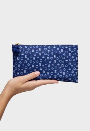 Mano sosteniendo una bolsa rectangular azul marino con símbolos de palos de cartas en azul claro y un tirador de cremallera con forma de trébol de cuatro hojas azul sobre un fondo gris.