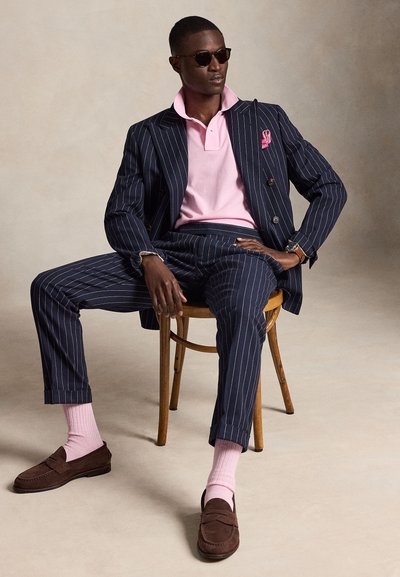 Uomo con abito blu navy a righe sottili, polo rosa, calzini rosa, mocassini marroni e occhiali da sole seduto su una sedia di legno in un ambiente neutro.