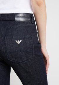 Jeans de denim oscuro con un ajuste elegante, que cuentan con una etiqueta de cuero en la cinturilla y un emblema de águila plateada en el bolsillo trasero.