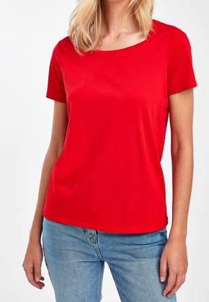 Vrouw draagt een effen rood T-shirt met korte mouwen en lichtblauwe spijkerbroek, getoond van schouders tot heupen tegen een witte achtergrond.